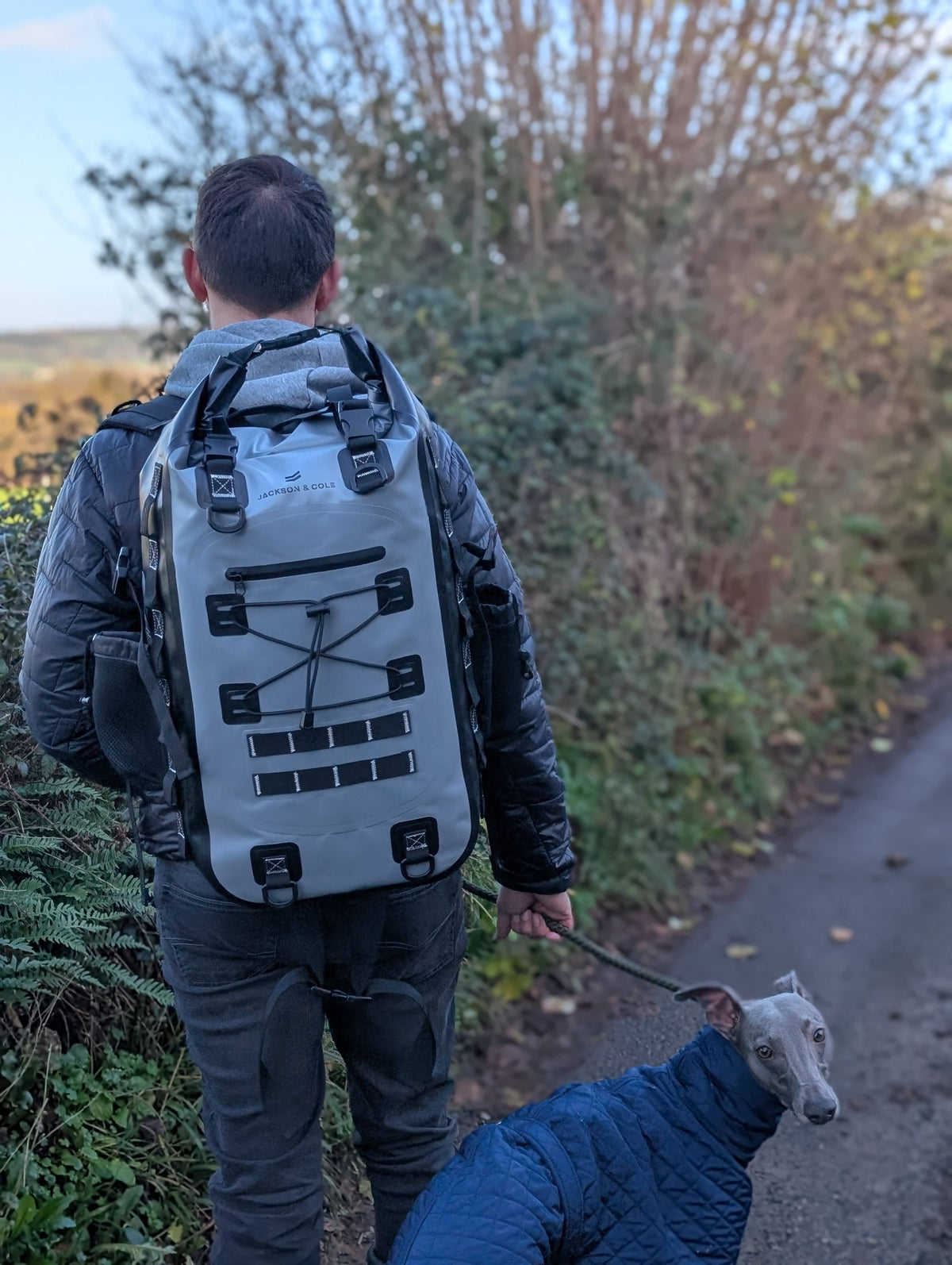 A man wearing a large gray waterproof backpack walks a grey dog on a paved path outdoors. The dog is wearing a blue quilted jacket.